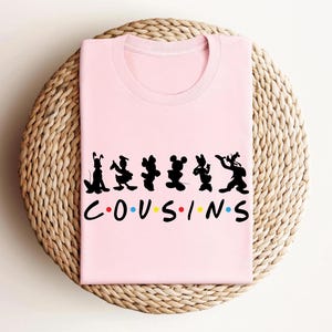 K&ouml;nnte beinhalten: Ein hellrosa T-Shirt mit einem schwarzen Silhouette-Design von Zeichentrickfiguren und dem Wort "COUSINS" in einer bunten Schriftart. Das Shirt liegt gefaltet auf einer gewebten, kreisf&ouml;rmigen Oberfl&auml;che.
