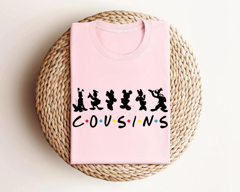 K&ouml;nnte beinhalten: Ein hellrosa T-Shirt mit einem schwarzen Silhouette-Design von Zeichentrickfiguren und dem Wort "COUSINS" in einer bunten Schriftart. Das Shirt liegt gefaltet auf einer gewebten, kreisf&ouml;rmigen Oberfl&auml;che.