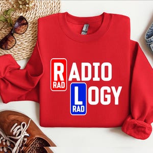 以下が含まれることがあります： 白い文字で「RADIOLOGY」と書かれた赤いスウェットシャツ。文字の両側に青い正方形で「RAD」と書かれている。