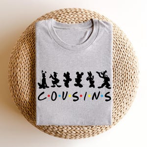 K&ouml;nnte beinhalten: Ein meliertes graues T-Shirt mit schwarzen Cartoon-Charakter-Silhouetten &uuml;ber dem Wort "COUSINS" in bunten Buchstaben. Das Shirt ist gefaltet und liegt auf einer gewebten, kreisf&ouml;rmigen Oberfl&auml;che.