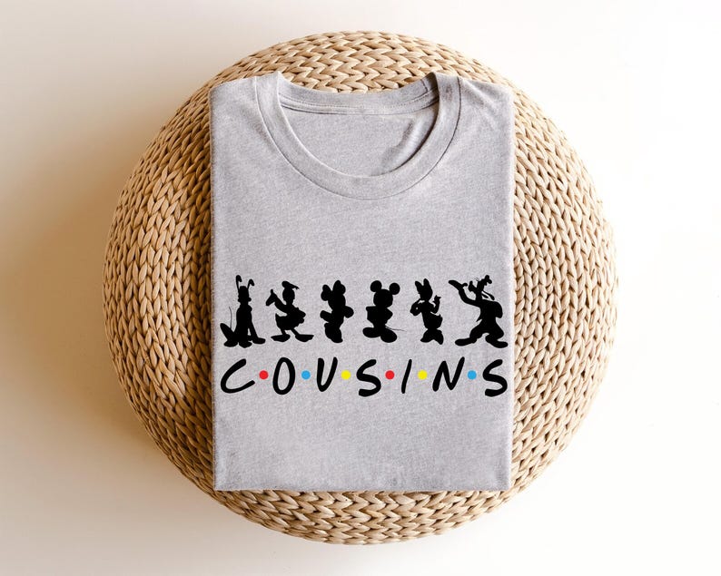 K&ouml;nnte beinhalten: Ein meliertes graues T-Shirt mit schwarzen Cartoon-Charakter-Silhouetten &uuml;ber dem Wort "COUSINS" in bunten Buchstaben. Das Shirt ist gefaltet und liegt auf einer gewebten, kreisf&ouml;rmigen Oberfl&auml;che.