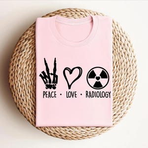 以下が含まれることがあります： 平和マーク、ハート、放射線記号が描かれたピンクのTシャツ。テキストは「Peace. Love. Radiology.」です