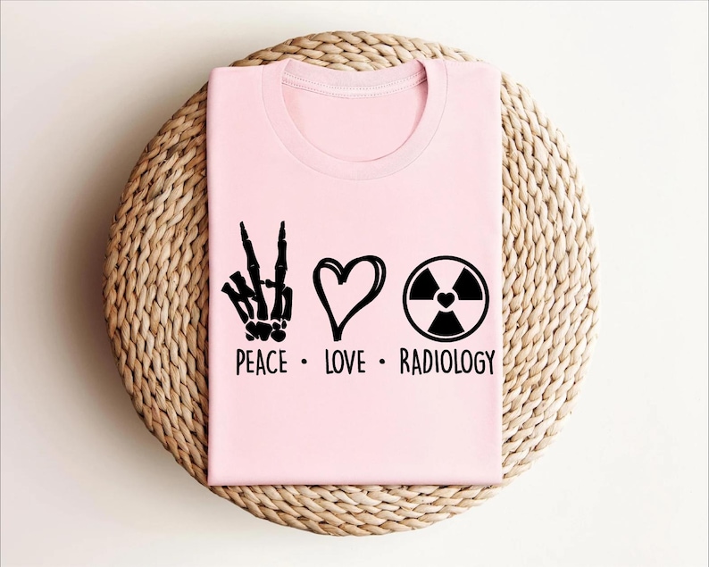 以下が含まれることがあります： 平和マーク、ハート、放射線記号が描かれたピンクのTシャツ。テキストは「Peace. Love. Radiology.」です