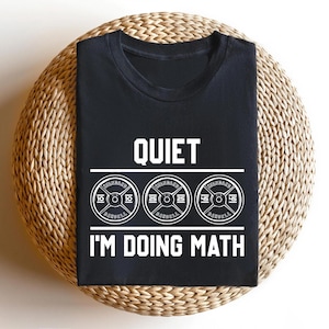 May include: Black t-shirt with the text "QUIET I'M DOING MATH" in white. Below the text are three barbell weight plates with the numbers 10, 25, and 45. The shirt is displayed on a round, woven, light brown surface.