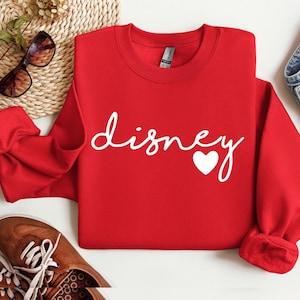 May include: A bright red long-sleeve crewneck sweatshirt featuring the word "disney" written in a white cursive script font, with a small white heart icon positioned at the end of the text. The garment has a ribbed collar and cuffs.