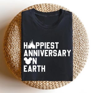 May include: Black t-shirt featuring the text "Happiest Anniversary on Earth" in white, with a castle and Mickey Mouse head design. The shirt is presented on a circular woven mat.