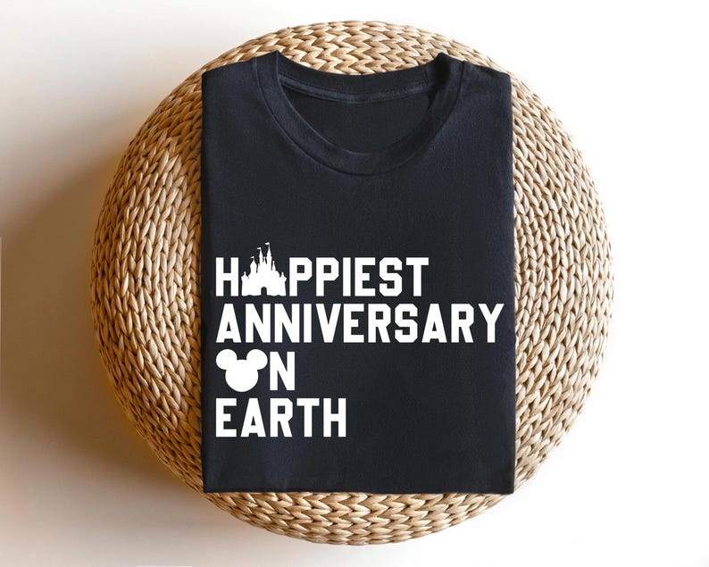 May include: Black t-shirt featuring the text "Happiest Anniversary on Earth" in white, with a castle and Mickey Mouse head design. The shirt is presented on a circular woven mat.