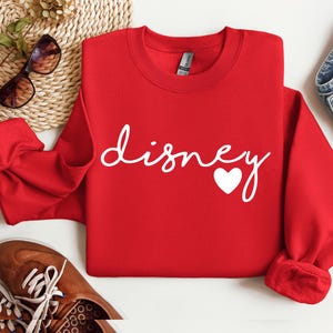 May include: A bright red long-sleeve crewneck sweatshirt featuring the word "disney" written in a white cursive script font, with a small white heart icon positioned at the end of the text. The garment has a ribbed collar and cuffs.