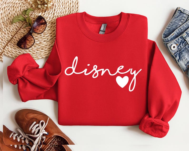 May include: A bright red long-sleeve crewneck sweatshirt featuring the word "disney" written in a white cursive script font, with a small white heart icon positioned at the end of the text. The garment has a ribbed collar and cuffs.