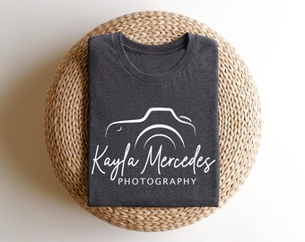 May include: Dark gray t-shirt with a white camera graphic and the text "Kayla Mercedes Photography". The shirt is folded neatly on a woven, circular surface. The design features a stylized camera outline.