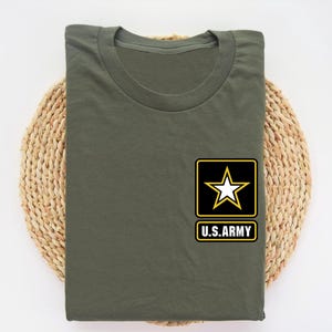 May include: Olive green t-shirt with a crew neck. The shirt features a black and yellow patch with a white star and the text "U.S. ARMY". The shirt is folded and placed on a woven, circular mat.