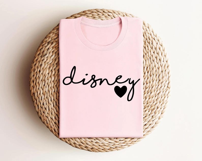 May include: A light pink t-shirt featuring the word "disney" written in a black cursive script font, with a solid black heart icon replacing the dot on the letter i. The garment has a classic crew neckline and short sleeves, presented folded.