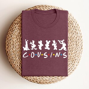 K&ouml;nnte beinhalten: Ein weinrotes T-Shirt mit einer wei&szlig;en Grafik mit Zeichentrickfiguren und dem Wort "COUSINS" in einer bunten Schriftart. Das Shirt ist gefaltet und auf einer gewebten, kreisf&ouml;rmigen Oberfl&auml;che platziert.