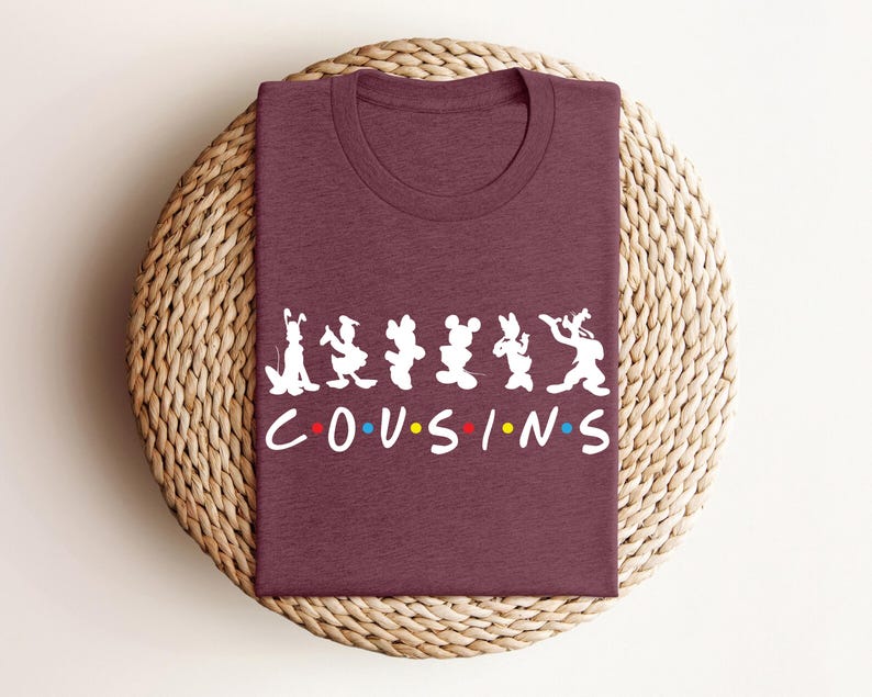 K&ouml;nnte beinhalten: Ein weinrotes T-Shirt mit einer wei&szlig;en Grafik mit Zeichentrickfiguren und dem Wort "COUSINS" in einer bunten Schriftart. Das Shirt ist gefaltet und auf einer gewebten, kreisf&ouml;rmigen Oberfl&auml;che platziert.