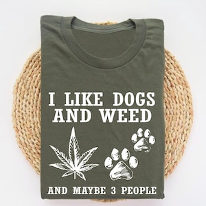 May include: Olive green t-shirt with the text "I LIKE DOGS AND WEED AND MAYBE 3 PEOPLE". The shirt features a white cannabis leaf and dog paw prints.