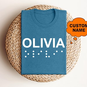 May include: A teal-colored t-shirt with the name "OLIVIA" in large white letters. Below the name are white dots representing Braille. An orange speech bubble with "CUSTOM NAME" is in the upper right corner.