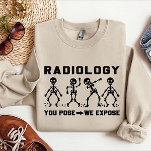 以下が含まれることがあります： ベージュのトレーナーに「RADIOLOGY YOU POSE - WE EXPOSE」という文字と、4つの骨格がさまざまなポーズをとっているグラフィックがプリントされています。