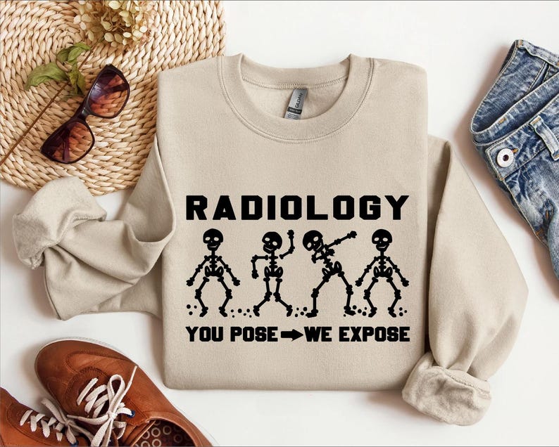 以下が含まれることがあります： ベージュのトレーナーに「RADIOLOGY YOU POSE - WE EXPOSE」という文字と、4つの骨格がさまざまなポーズをとっているグラフィックがプリントされています。