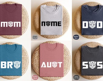 May include: Six different t-shirts with a Transformers robot head graphic and text that says "MOM", "NOME", "D0D", "BR0", "AUT", and "S0S". The shirts are in various colors, including maroon, heather gray, navy, teal, mauve, and dark gray.