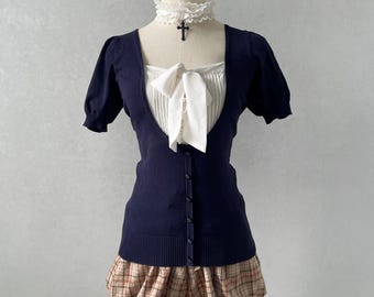 May include: A navy blue short-sleeved cardigan with a button-down front, worn over a white pleated blouse featuring a central bow. A white lace choker with a black cross pendant is worn at the neck. The outfit includes tan plaid bubble shorts.