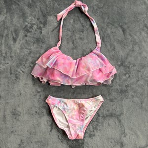 以下が含まれることがあります： ピンク、紫、白のタイダイ柄のツーピース水着セット。トップスはホルターネックで、フリルが施されています。ボトムはクラシックなビキニカットです。