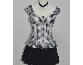 Puede incluir: Conjunto de top y falda a cuadros blancos y negros en un maniquí. El top presenta un escote cuadrado, ribete de encaje y cierre de botones. La falda es negra, en capas y con dobladillo con volantes. Un collarín y un collar negros completan el conjunto.