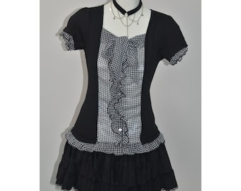 Puede incluir: Vestido de cuadros vichy en blanco y negro con un panel frontal con volantes y mangas cortas. El vestido presenta una falda negra escalonada y un collar gargantilla a juego con un colgante plateado. El vestido se exhibe en un maniquí.