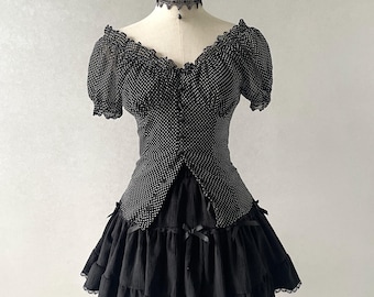 Puede incluir: Vestido negro con lunares blancos y gargantilla a juego, expuesto en un maniquí. El vestido presenta un escote con volantes, mangas cortas y una falda escalonada con ribete de encaje. Pequeños lazos negros adornan la falda.