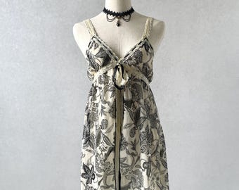 May include: A sleeveless mini dress featuring a cream base with a black floral print, thin lace-trimmed straps, a V-neckline with a black ribbon tie, and a black lace choker accessory. The dress has a loose, flowing silhouette and a hemline.