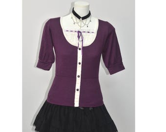 Puede incluir: Un suéter morado de manga corta con ribete de encaje blanco y botones negros. El conjunto incluye una falda negra en capas y un collar gargantilla negro con detalles plateados. La parte superior tiene una inserción de encaje blanco y una cinta morada.