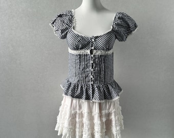 Puede incluir: Un vestido blanco con un corpiño de vichy blanco y negro y una falda de encaje blanco. El corpiño tiene un escote cuadrado y mangas cortas abullonadas. La falda es de capas y tiene un dobladillo con volantes.