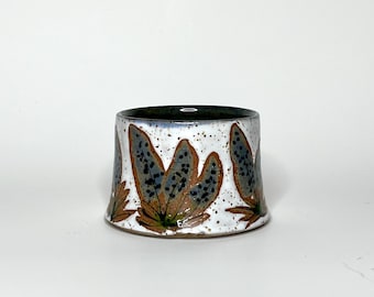 May include: A white ceramic cup with a brown rim and a pattern of blue and brown leaves around the middle.