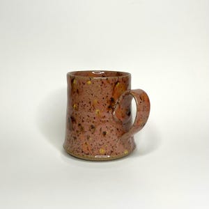 May include: A handmade ceramic mug with a speckled pink and brown glaze and gold flecks. The mug has a rounded body and a slightly curved handle.