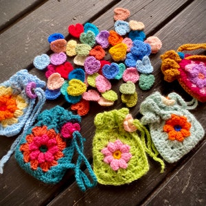 May include: A collection of crocheted drawstring pouches with colourful flower designs. The pouches are made with different coloured yarn, including blue, green, and pink. There are also a variety of crocheted hearts in different colours scattered around the pouches.
