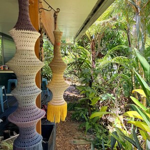 May include: Three handmade crochet lampshades hanging outdoors. The lampshades are in shades of brown, yellow, and blue, with a tiered design and fringe at the bottom. The background features lush green foliage.