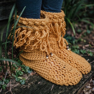 Può includere: Stivali all'uncinetto color giallo senape con frange e perline. Gli stivali sono fatti a mano con una cucitura strutturata e sembrano caldi e accoglienti. Sono su una superficie di legno, con vegetazione sullo sfondo.