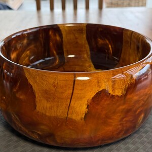 Puede incluir: Un cuenco de madera hecho a mano con un acabado brillante. El cuenco presenta una rica paleta de colores cálidos de marrones y ámbares, con patrones de vetas de madera visibles. El interior es liso y reflectante.