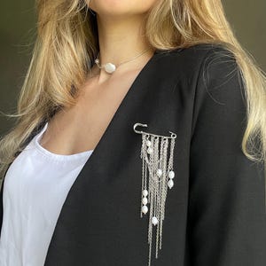Puede incluir: Un blazer negro con una parte superior blanca y una broche plateada. El broche presenta un diseño de imperdible grande con cadenas plateadas colgantes y perlas blancas. También se ve un collar de perlas.