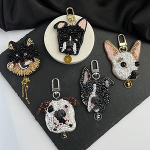 May include: Five dog-shaped keychains crafted with intricate beadwork in various colors including black, white, tan, and brown. Each keychain features a gold-toned metal clasp and a small circular charm with a letter attached to the bottom edge.