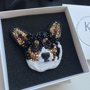 Puede incluir: Un broche de cuentas con forma de cabeza de perro Corgi, que presenta cuentas negras, blancas y doradas metálicas. El diseño incluye una nariz negra, ojos oscuros y orejas puntiagudas, todo meticulosamente elaborado con intrincados abalorios.