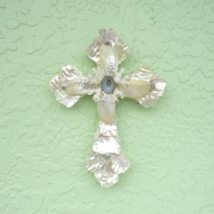 May include: A decorative cross made of iridescent seashells, with a central oval-shaped blue stone and coral-like accents. The cross is mounted on a textured, light green surface.