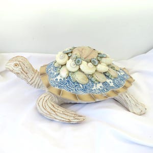 May include: A white ceramic turtle sculpture with a blue and gold patterned shell decorated with seashells. The turtle is positioned on a white surface.