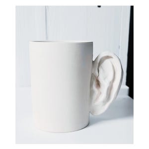 Puede incluir: Una taza de cerámica blanca mate con un asa escultural en forma de oreja humana. La taza tiene un cuerpo cilíndrico con un acabado liso y uniforme, y una oreja detallada y realista unida al lateral que sirve como asa funcional.