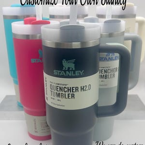 May include: A collection of customizable Stanley brand stainless steel tumblers with lids and straws. The tumblers are in various colors, including blue, pink, and gray. The text on the gray tumbler reads "The Flowstate™ Quencher H2.0 Tumbler 20 oz | 98 L All Day Hydration".