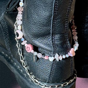 Puede incluir: Botas de cuero negro con una cadena plateada y una tobillera de cuentas. La tobillera presenta cuentas rosas, una cara sonriente, una luna creciente y dijes en forma de corazón. Las botas tienen una suela gruesa y están atadas.