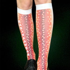 May include: Knee-high socks with a red and white spotted pattern, similar to deer markings. The socks have white cuffs and the text "DRINK WEAR" printed vertically. Black shoes are worn with the socks. The socks are worn on legs.