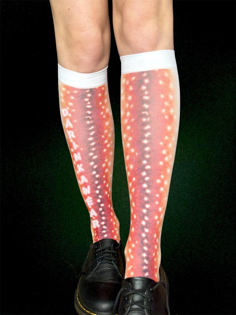 May include: Knee-high socks with a red and white spotted pattern, similar to deer markings. The socks have white cuffs and the text "DRINK WEAR" printed vertically. Black shoes are worn with the socks. The socks are worn on legs.