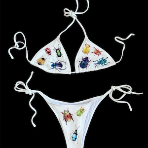 Op de afbeelding: Witte bikiniset met een driehoekige top en een laag uitgesneden broekje. De badkleding is versierd met kleurrijke insectenillustraties, waaronder kevers, in verschillende maten en kleuren. De bikini heeft witte bandjes.