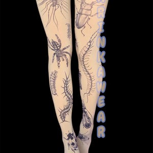 May include: Sheer beige tights with detailed black illustrations of various insects, including a spider, centipede, and beetle. The text "DARK KAWAII WEAR" is printed vertically on the right leg. The tights are worn against a black background.