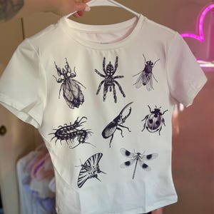 May include: White t-shirt featuring detailed black illustrations of various insects, including a spider, dragonfly, and beetle. The short-sleeved shirt is displayed on a white hanger.